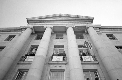 Sproul Hall with FSM signs hanging over the balconies. A student holds the American flag on the south balcony