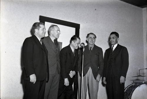 Five men standing in front of a microphone, one man bowing